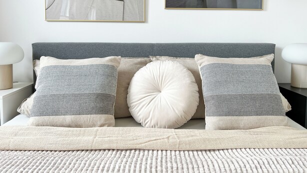 Een net opgemaakt bed met een grijze hoofdbord, twee gestreepte kussens en een beige plaid.