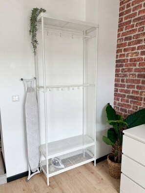 A white metal shelving unit with a mesh bottom shelf, a hanging plant, and a white dresser against a brick wall. - JUPAS° Soho Home – Studio with Parking (Trier)