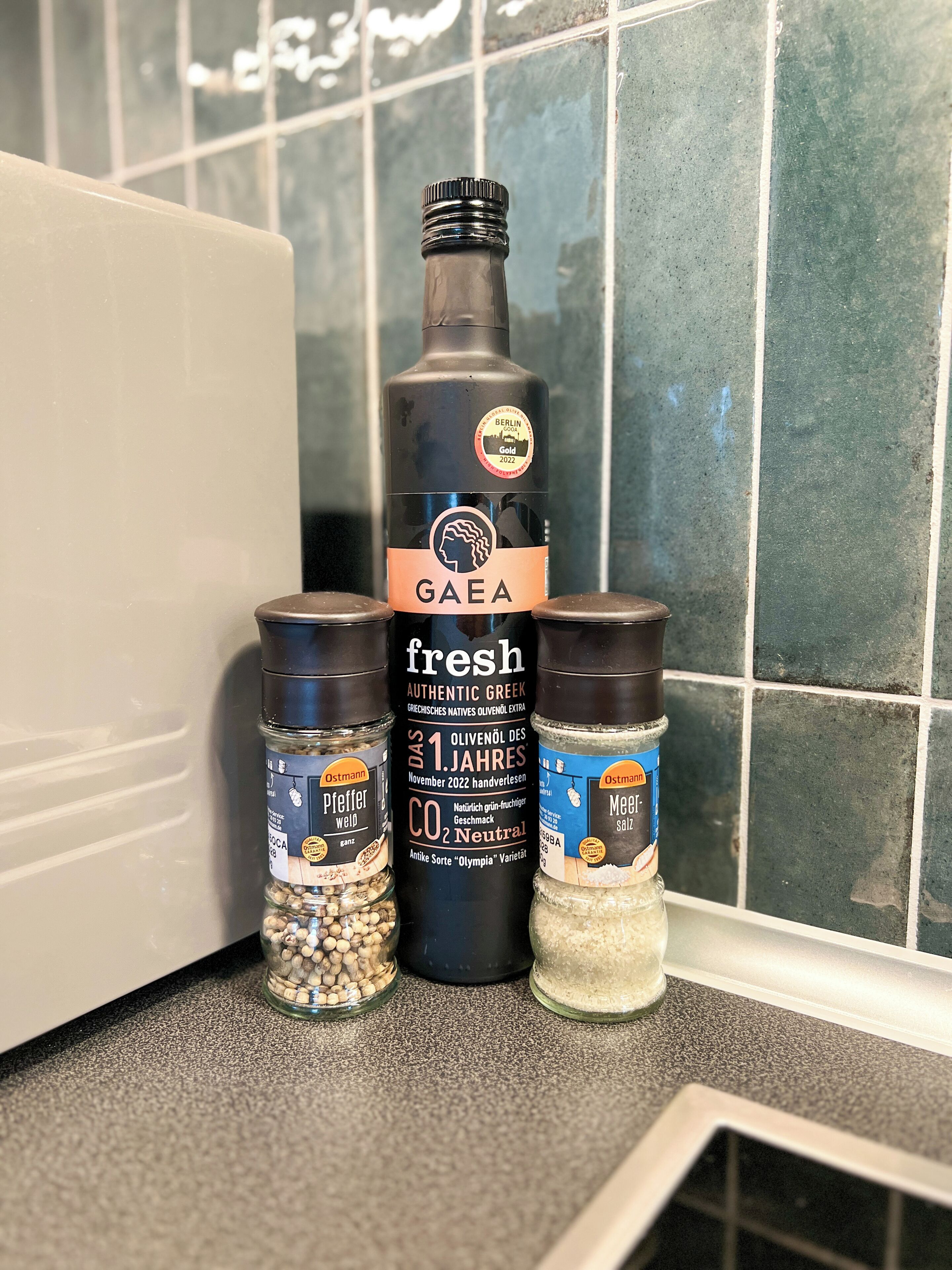 Una encimera de cocina con una botella de aceite de oliva griego auténtico y fresco GAEA, dos frascos de especias etiquetados como Pfeffer weid y Meer, y un recipiente de sal Ostmann.