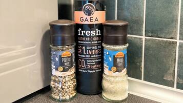 Una encimera de cocina con una botella de aceite de oliva griego auténtico y fresco GAEA, dos frascos de especias etiquetados como Pfeffer weid y Meer, y un recipiente de sal Ostmann.