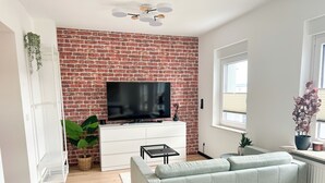 Een woonkamer met een bakstenen muur, een flatscreen-tv, een grijze bank, een wit kastje en een raam.