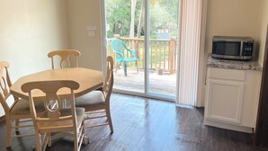 A dining area with a wooden table and chairs, a sliding glass door leading to a balcony with a view of greenery and a building, and a microwave on a cabinet.