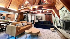 A spacious loft-style living area with a high ceiling, wooden beams, a mezzanine level, a fireplace, and a dining area with a view. - Riverfront A-frame Mt Hood! Skiing*Hiking*Hot tub (Rhododendron)