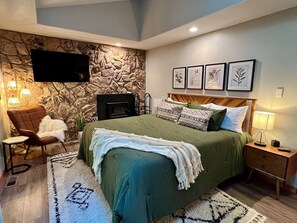 A bedroom with a stone wall, a bed with green bedding, a TV mounted on the wall, and framed pictures above the bed.
