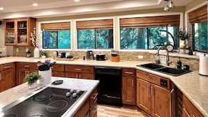 A modern kitchen with wooden cabinets, a central island with a stove, and large windows offering a view of the forest. - Riverfront A-frame Mt Hood! Skiing*Hiking*Hot tub (Rhododendron)