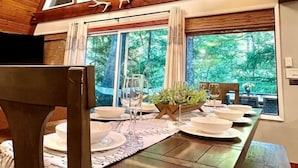 A dining area with a wooden table set for four, large windows with a forest view, and a chandelier. - Riverfront A-frame Mt Hood! Skiing*Hiking*Hot tub (Rhododendron)