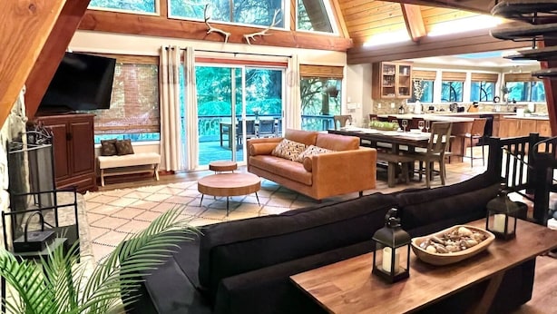A spacious living area with a large sofa, a wooden coffee table, and a dining table. There is a kitchen area in the background with a bar and seating. - Riverfront A-frame Mt Hood! Skiing*Hiking*Hot tub (Rhododendron)