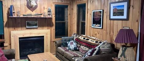 A cozy living room with a fireplace, wooden walls, and a sofa with patterned cushions.