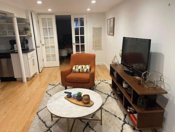 A living room with a sofa, a coffee table, a TV, and a kitchen area visible through an open door.