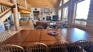 A spacious dining area with a large wooden table and chairs, a fireplace, and a television in a log cabin setting.