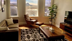 A living room with a sofa, armchair, coffee table, and TV stand. There are two windows with views of a mountain and a building. - The 1900 House - 1 Block From DT (Prescott)