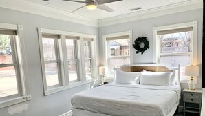 A bedroom with a large bed, two bedside tables, a ceiling fan, and a view of the outside through large windows. - The 1900 House - 1 Block From DT (Prescott)