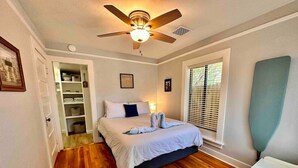 Une chambre à coucher comprenant un lit, un ventilateur de plafond, une fenêtre avec des stores, une porte et une étagère intégrée.