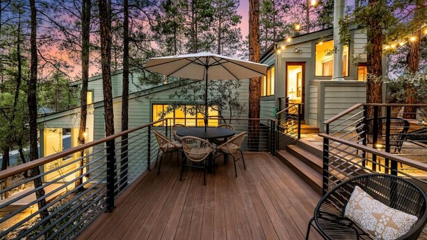 A wooden deck with a table and chairs, surrounded by tall trees and a building with a staircase. - Prescott Treehouse. Luxe 4BR/3Ba Home. Sleeps 10 (Prescott)
