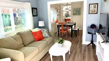 A living room with a beige sofa, a small white table with a plant, a dining area with a wooden table and chairs, and a kitchen area with a refrigerator and a sink.