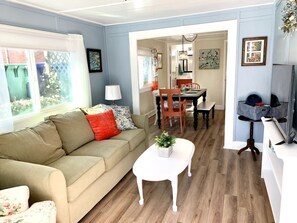 A living room with a beige sofa, a small white table with a plant, a dining area with a wooden table and chairs, and a kitchen area with a refrigerator and a sink. - Downtown Birdie / walk to whiskey row (Prescott)