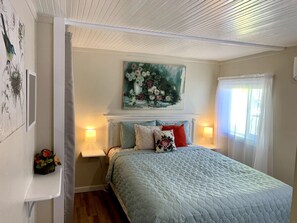 A bedroom with a bed, a painting above the bed, a nightstand, a lamp, and a window with curtains. - Downtown Birdie / walk to whiskey row (Prescott)
