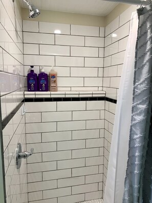 Zona de ducha con azulejos blancos, un cabezal de ducha y dos botellas de champú sobre una estantería.