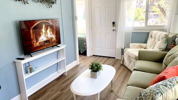 Una sala de estar con un televisor sobre un mueble blanco, una mesa de centro blanca con una planta, un sofá verde y una puerta blanca.