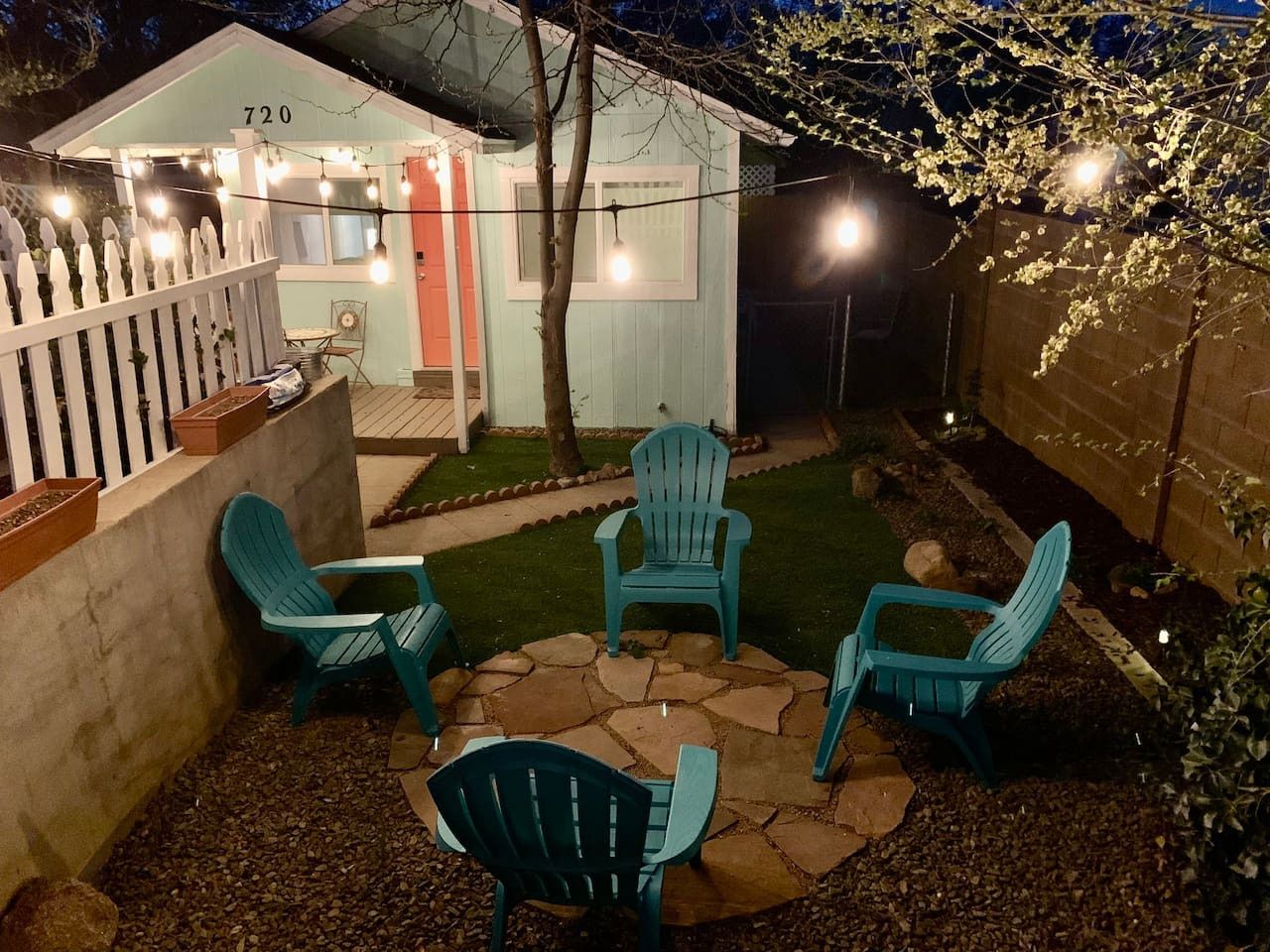 Un patio trasero con área de terraza que incluye cuatro sillas y una mesa, rodeado por una cerca blanca de piquete y un cobertizo pequeño con el número 720.