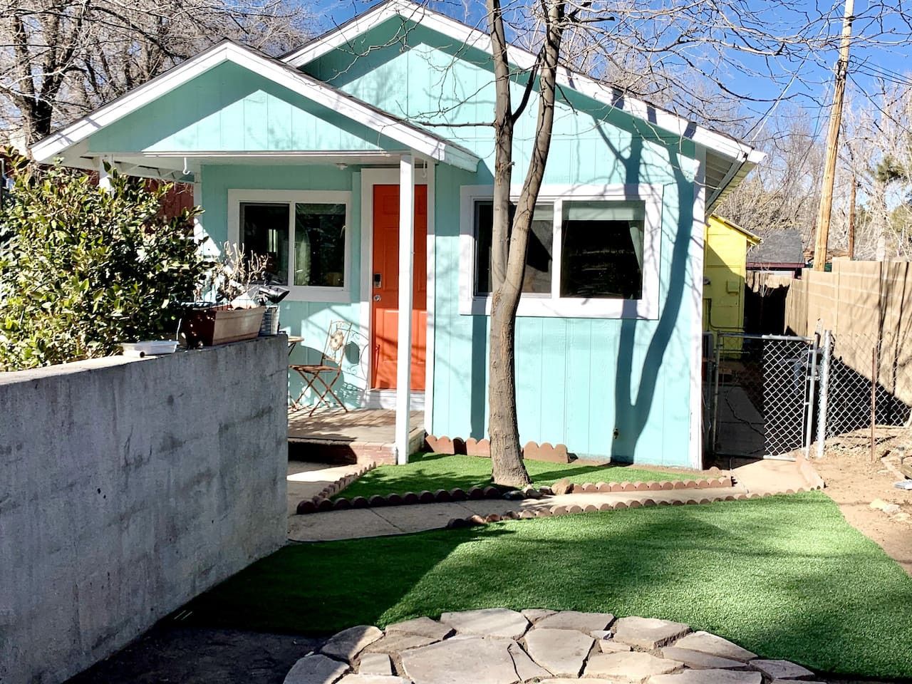 A single-story house with a teal exterior, a red door, and a small front yard with artificial grass and a stone pathway.
