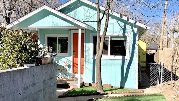 Une maison de plain-pied à la façade turquoise, avec une porte rouge et un petit jardin devant, doté d’une pelouse artificielle et d’un chemin en pierre.