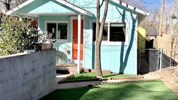 A single-story house with a teal exterior, a red door, and a small front yard with artificial grass and a stone pathway.