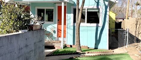 A single-story house with a teal exterior, a red door, and a small front yard with artificial grass and a stone pathway.