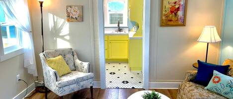 A living room with a wooden floor, a white armchair with a yellow cushion, a beige sofa with blue cushions, a white coffee table with a plant, and a kitchen visible through an open doorway.