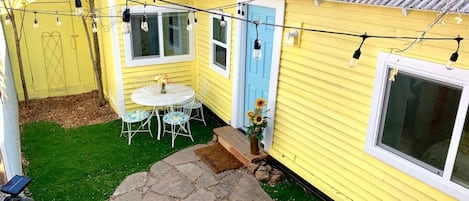 A yellow house with a blue door, a stone patio, a white table with chairs, and string lights.
