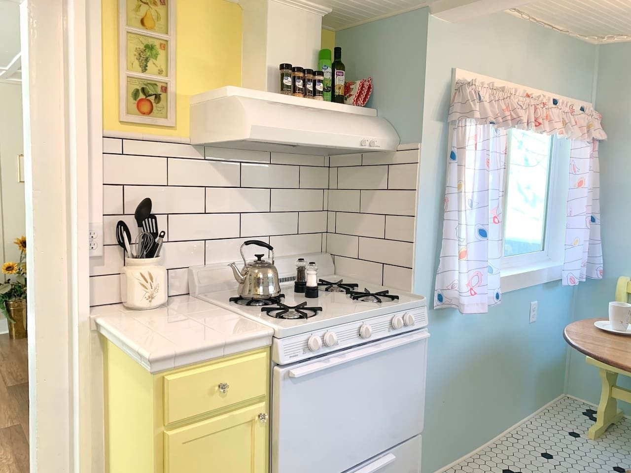 Une cuisine équipée d’une cuisinière à gaz, de carreaux blancs, d’un meuble jaune et d’une fenêtre ornée de rideaux à motifs.