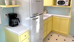 Eine kompakte Küche mit einem weißen Galanz-Kühlschrank, einer schwarzen Kaffeemaschine, gelben Schränken, einer weißen Arbeitsplatte und einer Mikrowelle auf einem gelben Regal.