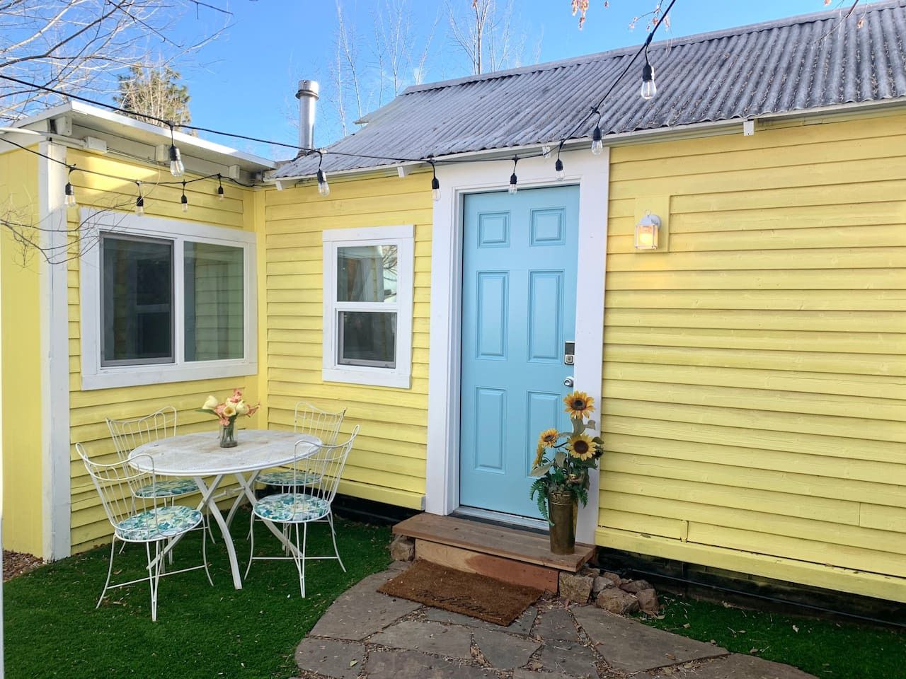Uma casa amarela com uma porta azul, um pequeno alpendre com mesa e cadeiras, e um vaso de flores.