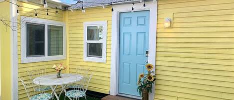 A yellow house with a blue door, a small porch with a table and chairs, and a flower vase.