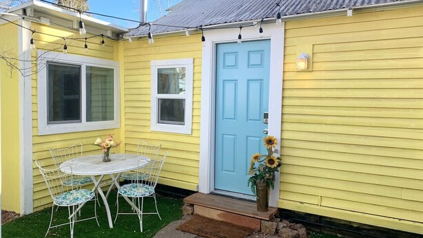 Ein gelbes Haus mit einer blauen Tür, einer kleinen Veranda mit Tisch und Stühlen sowie einer Blumenvase.