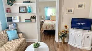 Ein Wohnzimmer mit einer Couch, einem Couchtisch mit Pflanze, einem Fernseher auf einem Schrank und einem Bett im Hintergrund.