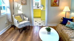 Ein Wohnzimmer mit Holzboden, einem weißen Sessel mit gelber Kissenauflage, einem beigen Sofa mit blauen Kissen, einem weißen Couchtisch mit Pflanze und einer durch einen offenen Durchgang sichtbaren Küche.