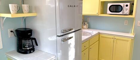 Uma cozinha compacta com uma geladeira Galanz branca, uma cafeteira preta, armários amarelos, bancada branca e micro-ondas em uma prateleira amarela.