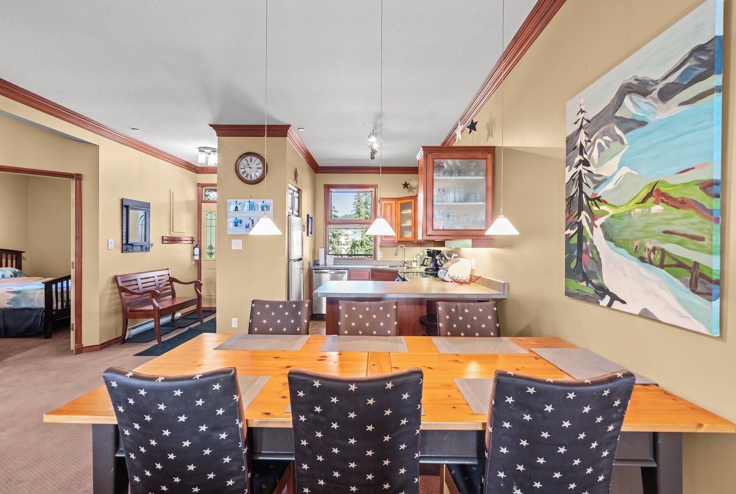 Un coin repas avec une table et des chaises en bois, une cuisine à l’arrière-plan et un tableau accroché au mur.