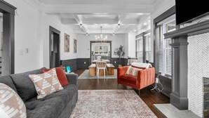 A spacious living room with a dark gray sofa, a red armchair, and a patterned rug. There is a dining area in the background with a table and chairs. - Walnut Charming Modern 5BR Home Walkable Kings (Kansas City)