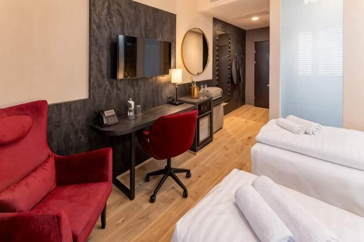 Ein Hotelzimmer mit Schreibtisch, einem roten Sessel, einem Bett mit weißer Bettwäsche, einem Spiegel und einem Fernseher.