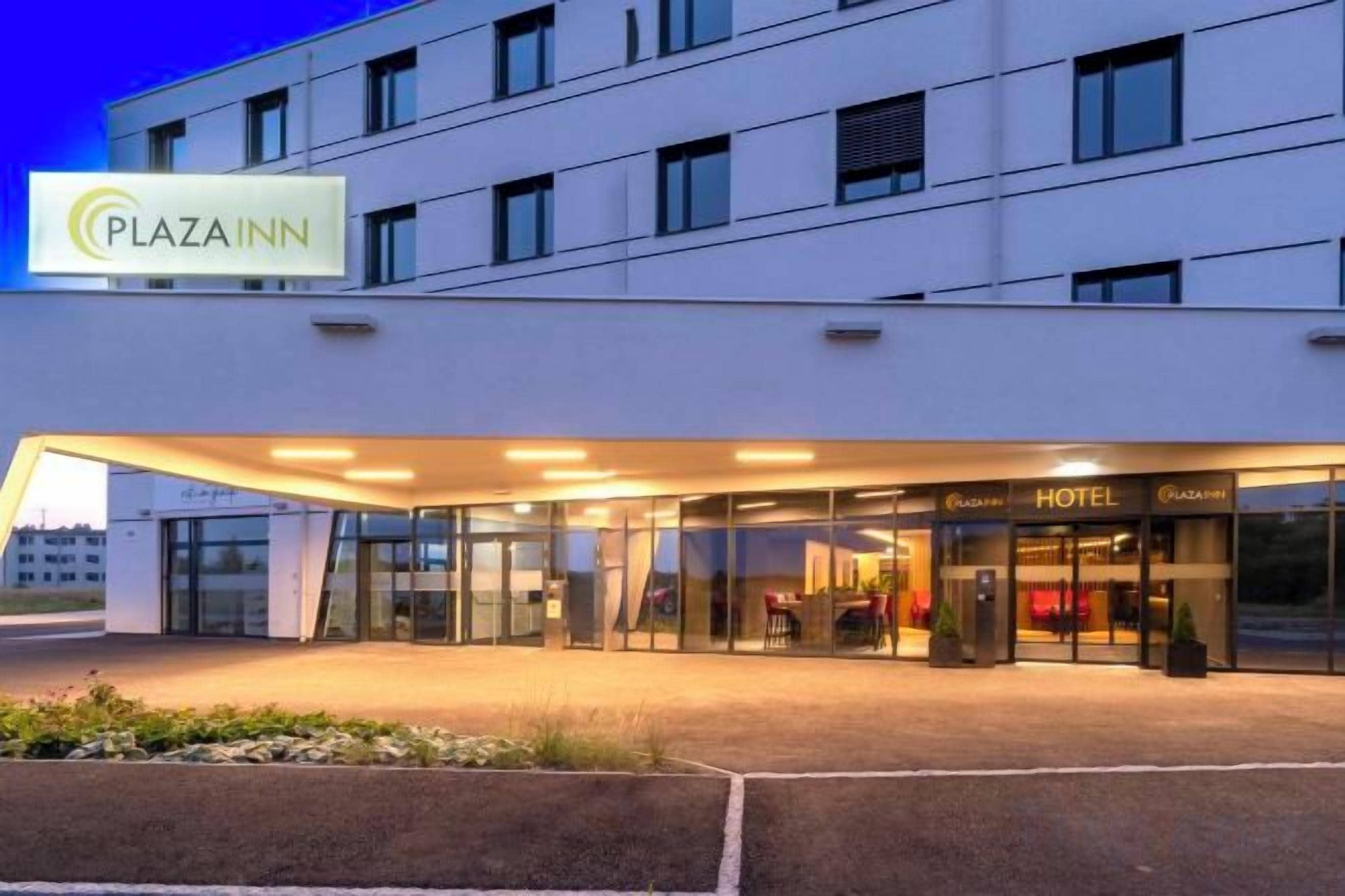 Une entrée d’hôtel moderne avec une façade en verre, un espace d’entrée abrité et une enseigne indiquant qu’il s’agit d’un hôtel Plaza Inn.