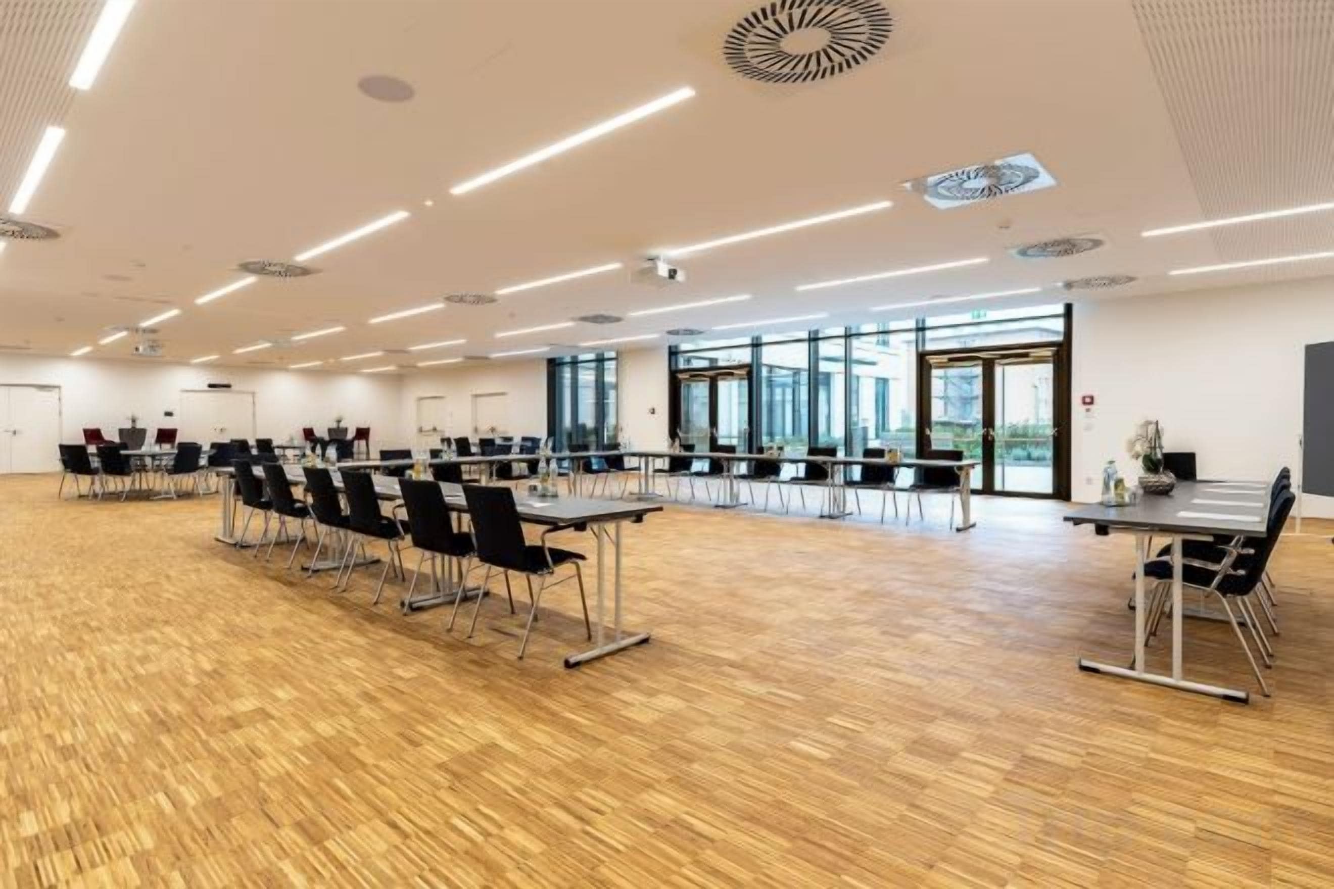 Un salón de conferencias con filas de mesas y sillas, ventanas amplias y suelo de madera.