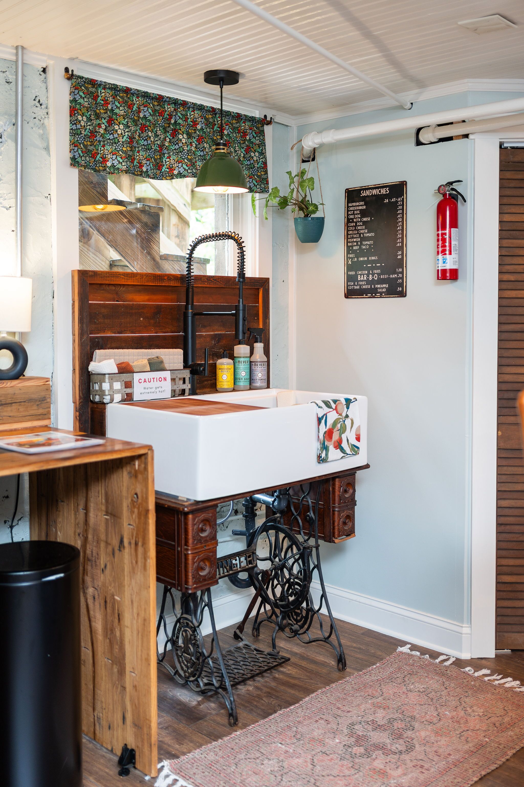 A vintage-style bathroom with a wooden vanity, a sink, and a mirror. There is a floral-patterned shower curtain, a hanging plant, and a fire extinguisher mounted on the wall.