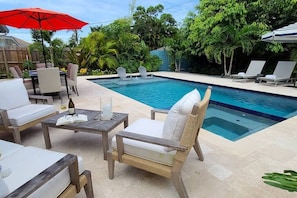 A poolside area with lounge chairs, a table with a bottle and book, and a red umbrella. - 677 Mod Beach Bliss | Heated Pool & Spa (Naples)