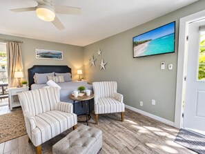 A hotel room with a bed, two armchairs, a small table, a ceiling fan, a painting of a beach, and a door with a doormat. - 677 Beach Break ·Beautiful 3/3 Home w/ Heated Pool (Naples)