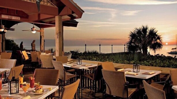 An outdoor dining area with tables and chairs, overlooking a body of water and palm trees during sunset. - 677 Beach Break ·Beautiful 3/3 Home w/ Heated Pool (Naples)