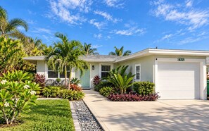 A residential house with a garage, surrounded by lush tropical plants and a clear blue sky. - 677 Beach Break ·Beautiful 3/3 Home w/ Heated Pool (Naples)