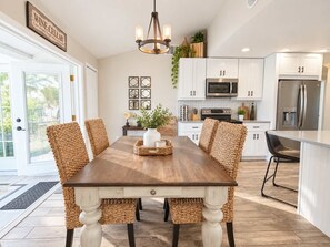 A modern kitchen with white cabinets, stainless steel appliances, and a dining area with a wooden table and woven chairs. - 677 Beach Break ·Beautiful 3/3 Home w/ Heated Pool (Naples)
