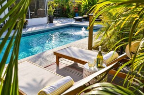 A pool area with lounge chairs, a table with a bottle and glass, and surrounding greenery. - 677 Beach Break ·Beautiful 3/3 Home w/ Heated Pool (Naples)